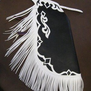 May include: A black and white leather saddle flap with a decorative white fringe and cut-out design. The flap has a white leather strap and buckle. The design includes geometric shapes and curved lines.