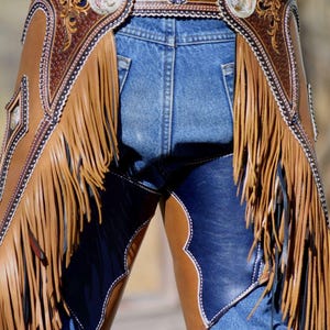 Op de afbeelding: Close-up van op maat gemaakte leren chaps met ingewikkelde details en franjes. De chaps zijn bruin en blauw, met zilveren accenten op de riem en decoratieve elementen. De tekst "Denice Langley Custom" is zichtbaar bovenaan.