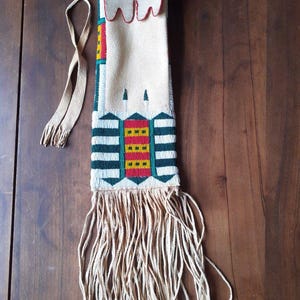 Op de afbeelding: Een crèmekleurig, met kralen bezet zakje met lange franjes. Het zakje heeft een geometrisch ontwerp met rode, gele, groene en zwarte kralen. Een lange, crèmekleurige riem is aan de bovenkant bevestigd. Het zakje wordt op een houten oppervlak gepresenteerd.