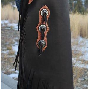 Op de afbeelding: Zwarte leren chaps met een bruine, getextureerde riem en decoratieve zilveren concho's. De chaps hebben een gefranjerde onderkant en worden over een spijkerbroek gedragen. Western wear.