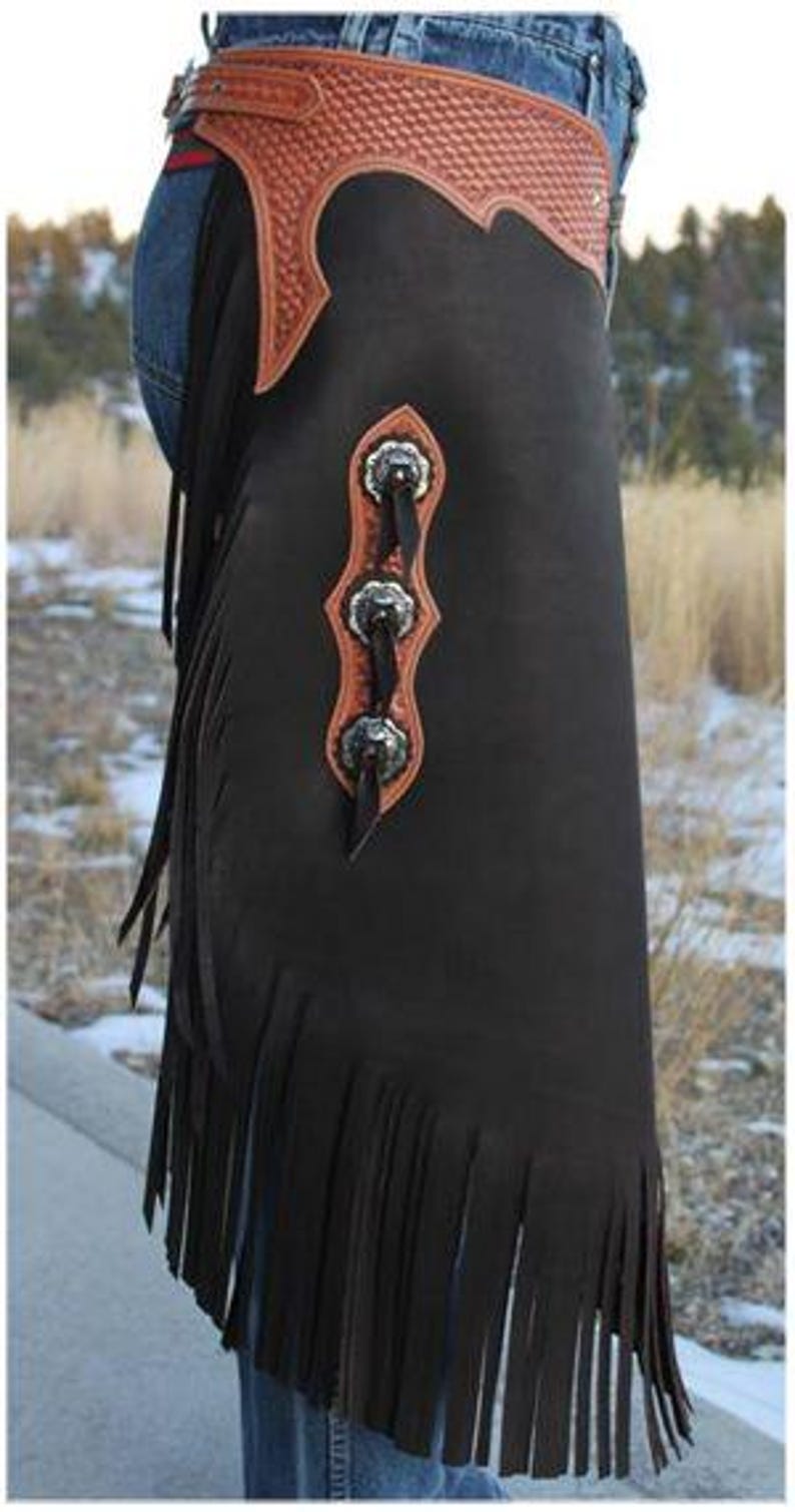 Op de afbeelding: Zwarte leren chaps met een bruine, getextureerde riem en decoratieve zilveren concho's. De chaps hebben een gefranjerde onderkant en worden over een spijkerbroek gedragen. Western wear.