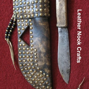 Puede incluir: Un juego artesanal de cuchillo y funda. La funda de cuero marrón oscuro está adornada con tachuelas de latón y un recorte decorativo. El cuchillo tiene un mango de madera y una hoja de metal desgastada. El texto "Leather Nook Crafts" es visible.