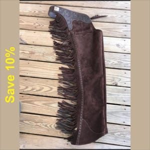 May include: Brown suede chaps with silver accents and fringe detailing. The chaps are a dark brown colour, designed for riding. The image shows the chaps laid out on a wooden surface.