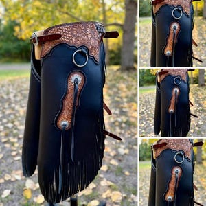 May include: Black leather chaps with brown floral tooled leather detailing. The chaps have a sunflower design on the yoke, silver hardware, and fringe along the bottom. Adjustable straps and a metal ring are also visible.
