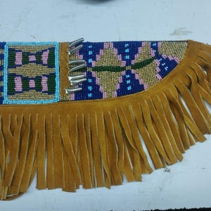 Pode incluir: Uma clutch de camurça castanha com detalhes de contas e franjas. A clutch apresenta uma seção de contas retangular com contas azuis, rosa e douradas. Detalhes metálicos prateados estão presentes. A borda inferior tem franjas longas e fluidas.