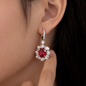 Può includere: Un orecchino d'argento con una grande gemma rossa rotonda al centro, circondata da gemme chiare più piccole. L'orecchino è a goccia, con un cerchio anch'esso incastonato con piccole gemme chiare. L'orecchino è mostrato sull'orecchio di una persona.