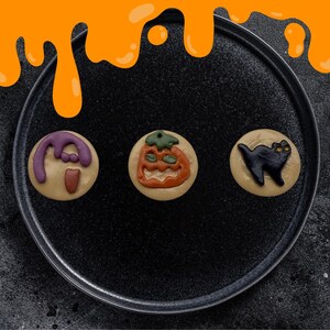 Może przedstawiać: Trzy ciasteczka o tematyce Halloween na czarnym talerzu. Ciasteczka przedstawiają fioletowego ducha, pomarańczową dynię i czarnego kota. Talerz stoi na ciemnej powierzchni, z pomarańczowymi kroplami w tle.