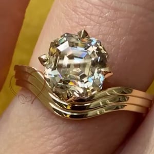 May include: A gold engagement ring with a large, faceted diamond center stone. The ring has a unique, curved band design. The diamond sparkles, reflecting light. The ring is set on a finger, showcasing its elegant design.