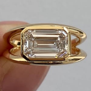 May include: A gold ring with an emerald-cut diamond. The diamond is set in a bezel setting, and the ring has a distinctive, open design. The ring is a bright gold colour and is shown against a neutral background.
