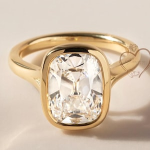 May include: A gold ring with a large, square-cut, clear gemstone. The gemstone is set in a bezel setting, and the band is a simple, polished gold. The ring is displayed on a neutral background, highlighting its elegant design.