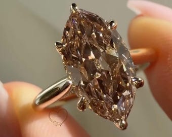 以下が含まれることがあります： シャンパンカラーのマーキスカットの宝石が施された、磨き上げられた金属バンドの指輪。石は温かみのある茶色と金色の色調のファセット面を持ち、6本の爪で固定されています。画像上部にHONEY CHAMPAGNEというテキストが表示されています。