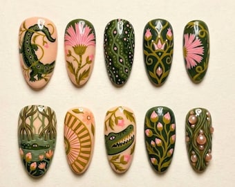 May include: A set of ten almond-shaped press-on nails featuring intricate hand-painted designs in shades of olive green, soft pink, and cream. The artwork includes detailed illustrations of crocodiles, swamp flora, floral patterns, and small pearl accents.