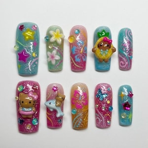 May include: A set of ten artificial nails with a variety of designs. The nails feature glitter, floral, and character embellishments in shades of pink, blue, and yellow. Some nails have 3D elements like flowers and stars.