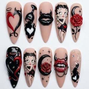 May include: A set of ten long, almond-shaped artificial nails with various designs. The designs include a devil heart, a wine glass, a heart, Betty Boop, a crescent moon with a rose, and lips with horns. The nails are painted in black, red, and nude colors.