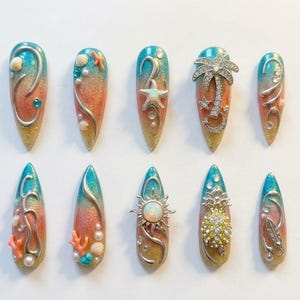May include: A set of ten long, almond-shaped artificial nails with a gradient design of blue, orange, and gold. Each nail features intricate embellishments, including seashells, starfish, coral, and silver swirls, creating a beach-themed aesthetic.