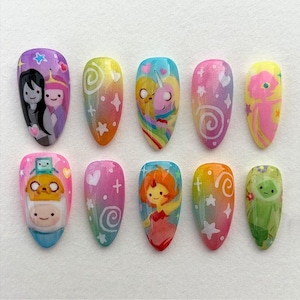 Pode incluir: Dez unhas postiças em formato amêndoa com desenhos pintados à mão inspirados em personagens de Adventure Time. O conjunto inclui ilustrações de Finn, Jake, Princesa Jujuba, Marceline, Princesa de Fogo e BMO, acentuadas com estrelas, corações e espirais.