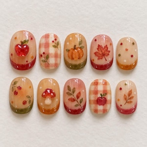 Op de afbeelding: Een set van tien ovale press-on nagels met herfstthema's, waaronder een rode appel, oranje pompoen, rode paddenstoel, esdoornblad en ruitpatronen in tinten rood, oranje en crème met kleine geschilderde stippen en bladeren.