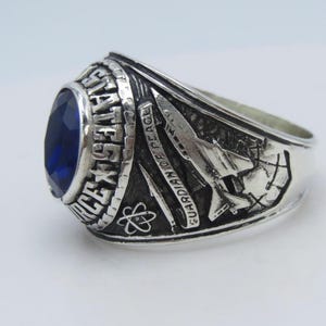 May include: Silver-toned ring with a large, oval, blue gemstone. The ring features detailed engravings, including text that reads "AIR FORCE" and "GUARDIAN OF PEACE." The design includes symbols and imagery associated with the Air Force.