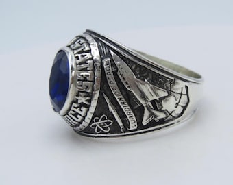 May include: Silver-toned ring with a large, oval, blue gemstone. The ring features detailed engravings, including text that reads "AIR FORCE" and "GUARDIAN OF PEACE." The design includes symbols and imagery associated with the Air Force.