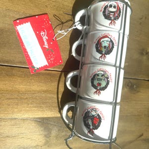 May include: A set of four white ceramic mugs stacked in a silver metal rack. Each mug features a graphic of characters from The Nightmare Before Christmas and the text Making Christmas Fright. A red Disney tag is attached, noting 6 fl oz each.