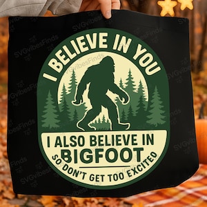 Op de afbeelding: Zwarte draagtas met een cirkelvormig ontwerp met een Bigfoot-silhouet voor een bos. De tekst luidt "I BELIEVE IN YOU" en "I ALSO BELIEVE IN BIGFOOT SO DON'T GET TOO EXCITED".