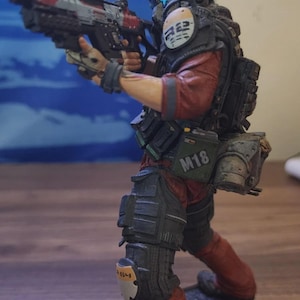 Puede incluir: Una figura de acción detallada de un soldado con uniforme táctico rojo, que lleva un casco con visera azul. La figura sostiene un rifle y presenta un parche en el hombro con texto, una bolsa M18 verde en el cinturón y rodilleras con marcas.