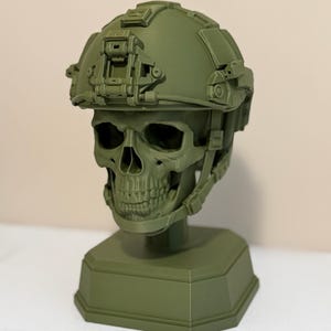 May include: An olive green monochromatic sculpture of a human skull wearing a tactical combat helmet. The helmet features a front-mounted night vision shroud, side rails, and a chin strap system. The entire piece is mounted on a solid octagonal base.