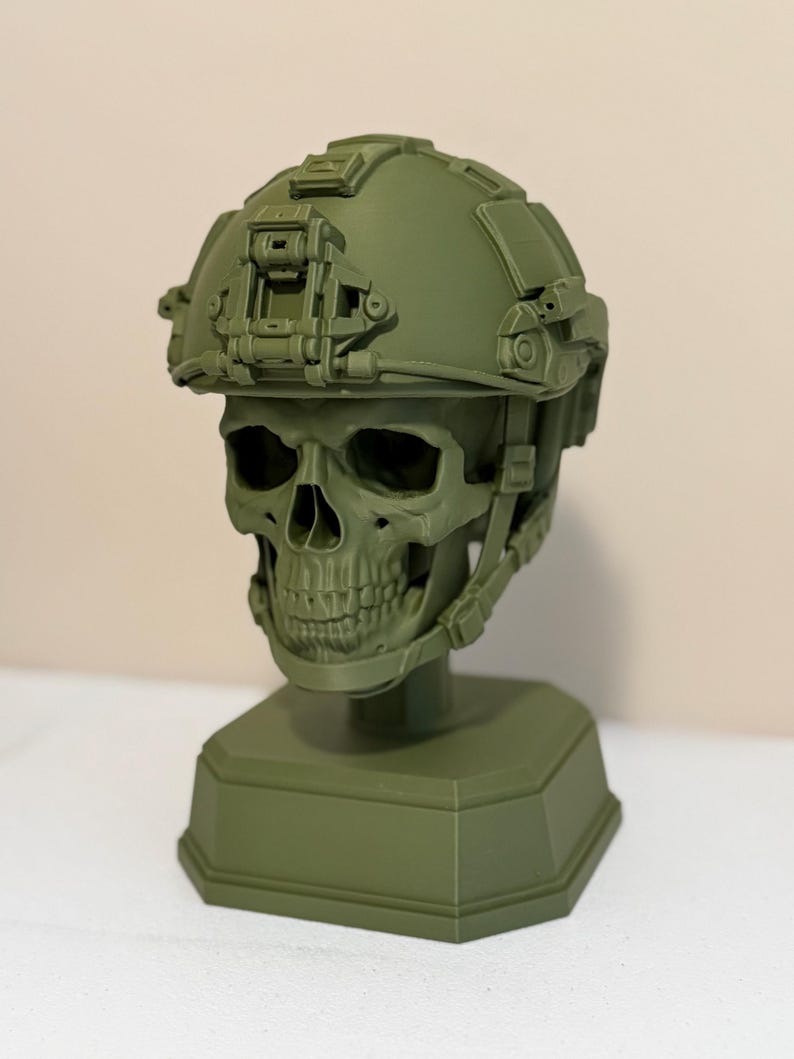 May include: An olive green monochromatic sculpture of a human skull wearing a tactical combat helmet. The helmet features a front-mounted night vision shroud, side rails, and a chin strap system. The entire piece is mounted on a solid octagonal base.