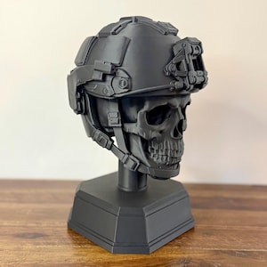May include: A matte black skull sculpture wearing a tactical combat helmet with an integrated mounting bracket on the front. The skull is mounted on a solid hexagonal base, featuring detailed bone structure and realistic helmet straps and hardware.