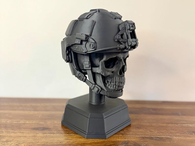 May include: A matte black skull sculpture wearing a tactical combat helmet with an integrated mounting bracket on the front. The skull is mounted on a solid hexagonal base, featuring detailed bone structure and realistic helmet straps and hardware.