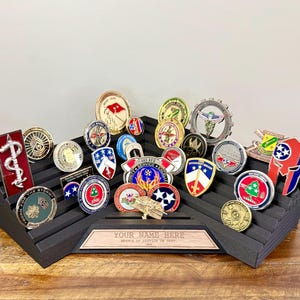 May include: A black tiered display stand holding a collection of various military challenge coins. The stand features a central wooden plaque with the engraved text "YOUR NAME HERE" and "BRANCH OF SERVICE OR DEPT." The coins vary in shape, color, and design.