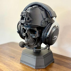 May include: A matte black skull sculpture wearing a tactical helmet and black HyperX gaming headset with a boom microphone. The skull is mounted on a solid black hexagonal base, creating a display stand for audio equipment.