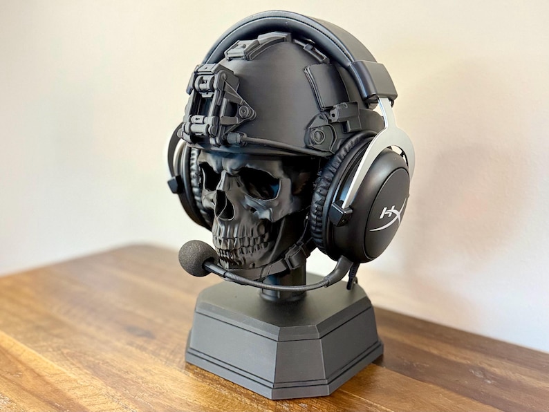May include: A matte black skull sculpture wearing a tactical helmet and black HyperX gaming headset with a boom microphone. The skull is mounted on a solid black hexagonal base, creating a display stand for audio equipment.