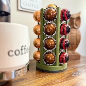May include: A green, cylindrical coffee pod holder with multiple slots, holding various coffee pods in gold, brown, and red striped designs. A white coffee mug with the word "coffee" is visible on the left. A wooden owl statue is in the background.