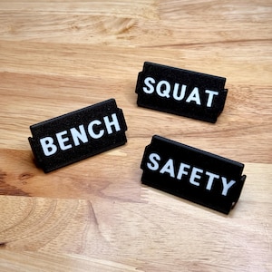 May include: Three black rectangular signs with white text that read "BENCH", "SQUAT", and "SAFETY". The signs are on a light brown wooden surface. The signs are likely for a gym or fitness setting.