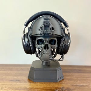 May include: A matte black skull sculpture wearing a tactical combat helmet and black over-ear headphones with a microphone boom. The skull is mounted on a solid, hexagonal black base, creating a functional display stand for gaming headsets.