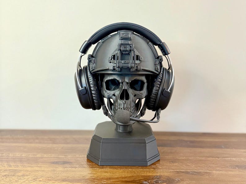 May include: A matte black skull sculpture wearing a tactical combat helmet and black over-ear headphones with a microphone boom. The skull is mounted on a solid, hexagonal black base, creating a functional display stand for gaming headsets.