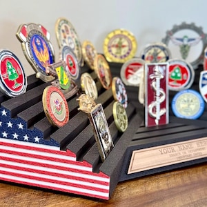 May include: A tiered black display stand for challenge coins featuring a printed American flag pattern on the side. The stand includes horizontal grooves for holding multiple circular and rectangular coins and a wooden plaque with the text YOUR NAME HERE.