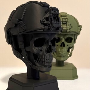 May include: Two skull busts wearing tactical helmets, one in matte black and one in olive green. Each skull is mounted on a hexagonal base and features a detailed helmet with a front-mounted night vision goggle shroud and side rail accessories.