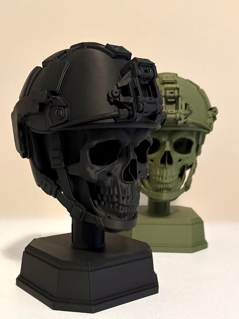 May include: Two skull busts wearing tactical helmets, one in matte black and one in olive green. Each skull is mounted on a hexagonal base and features a detailed helmet with a front-mounted night vision goggle shroud and side rail accessories.