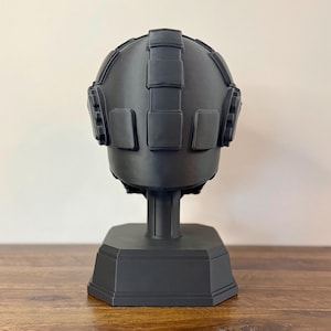 May include: A matte black helmet display stand featuring a rounded dome shape with raised rectangular panel details across the top and sides. The helmet is mounted on a central vertical support column attached to a sturdy, octagonal base.