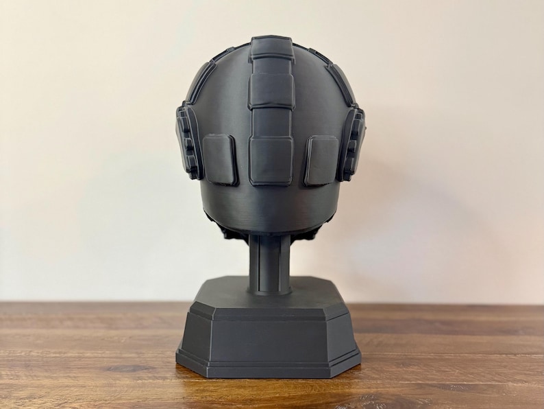 May include: A matte black helmet display stand featuring a rounded dome shape with raised rectangular panel details across the top and sides. The helmet is mounted on a central vertical support column attached to a sturdy, octagonal base.
