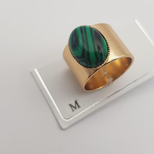 Peut inclure: Une large bague en métal doré ornée d'une grande pierre ovale aux rayures vertes et noires, sertie dans un chaton perlé. La bague est posée sur un support blanc marqué de la lettre M, révélant une texture brossée sur l'anneau.