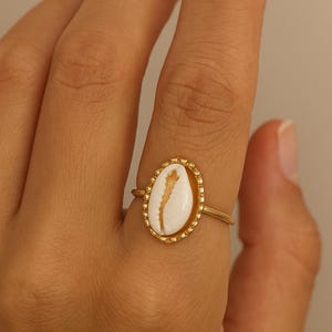 May include: A gold ring featuring a central white cowrie shell set within an oval frame. The metal band is thin and polished, while the shell is surrounded by a delicate beaded edge detail that adds texture to the overall design of the jewellery piece.
