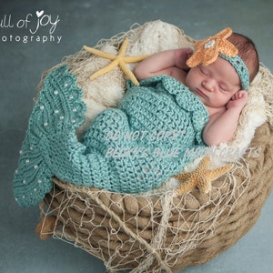 May include: A newborn baby wearing a crocheted teal mermaid costume and a crocheted orange flower headband, sleeping in a woven basket with a starfish and netting.