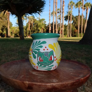 May include: A white ceramic pot with a green rim, featuring a hand-painted green creature face with red eyes in the center, surrounded by yellow and green leaf-like patterns and small red dots, resting on a round wooden surface.