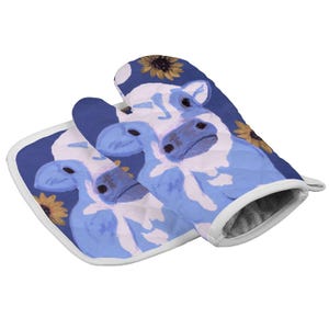 Op de afbeelding: Een gewatteerde ovenwant en een bijpassende vierkante pannenlap met een blauw-wit koeienpatroon op een donkerblauwe achtergrond, geaccentueerd met gele zonnebloemen. Beide items zijn afgewerkt met een witte stoffen bies langs de randen.
