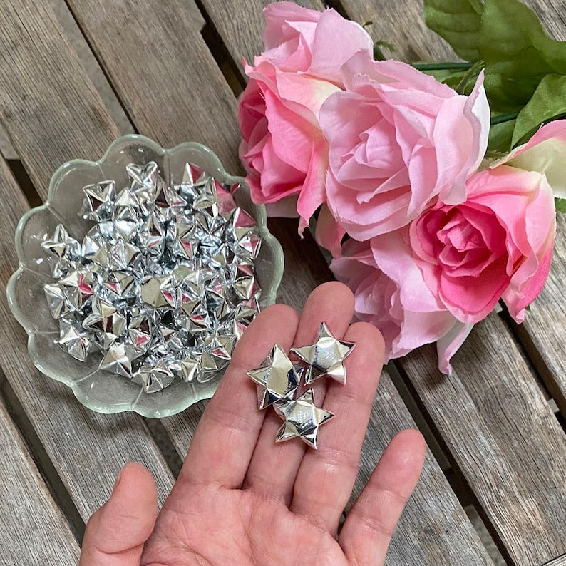 Puede incluir: Un bol lleno de estrellas de origami plateadas, con una mano sosteniendo algunas estrellas plateadas m&aacute;s. Las estrellas est&aacute;n hechas de papel met&aacute;lico y tienen un acabado brillante.