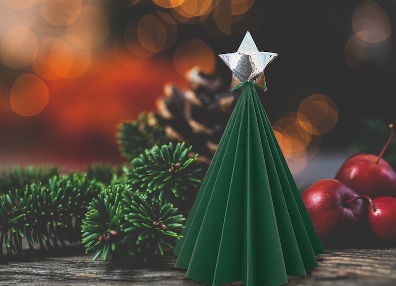 Puede incluir: Un &aacute;rbol de Navidad de papel verde con una estrella plateada en la parte superior. El &aacute;rbol est&aacute; sobre una superficie de madera con ramas de pino y manzanas rojas.