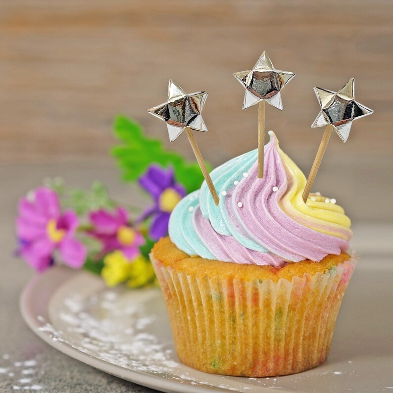Puede incluir: Un cupcake colorido con glaseado azul pastel y morado cubierto con tres adornos en forma de estrella plateados. El cupcake est&aacute; en un plato blanco con un fondo de madera marr&oacute;n claro.